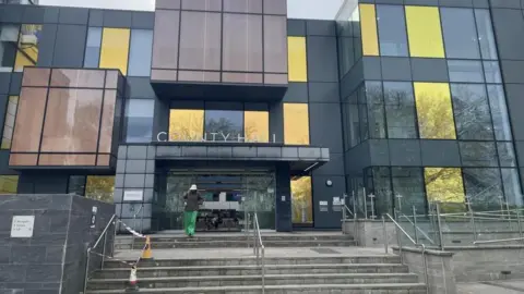 Peter Davison The front entrance of County Hall in Trowbridge with a person wearing a white hat and green trousers walking towards the entrance.