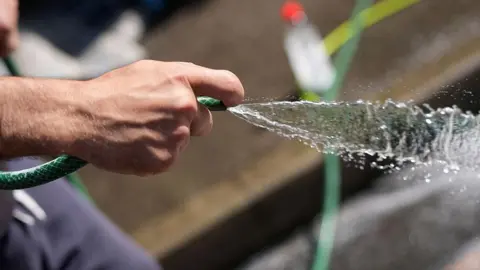 A close-up picture of a hand using a green hosepipe. Water is spraying out of the pipe.
