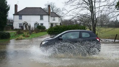 PA Media A car passes through a flooded road with a white house in the background.