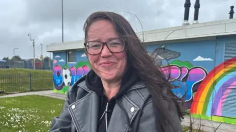 Zoe, who has long brown hair and is wearing a leather jacket and glasses, smiles
