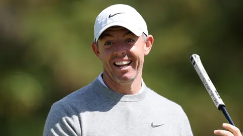 Rory McIlroy smiles past the camera as he holds a golf putter. He is wearing a grey sweatshirt and a white cap.