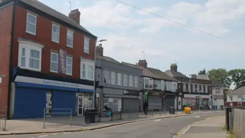 A road in Hull showing a row of high street shops with shutters closed. There is a path and a road in front of the shops. The street is empty and the sky is cloudy.