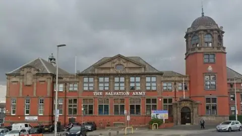 Google A large red brick building with a sign saying 'The Salvation Army'. Some cars are parked in front.