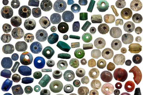 Anna Giecco Dozens of old beads, of various shapes and sizes, are laid out on a plain white background and arranged by colours. It goes from dark blue in the top left hand corner to red beads at the bottom right - gradually going through shades of green, yellow and pink on the way.