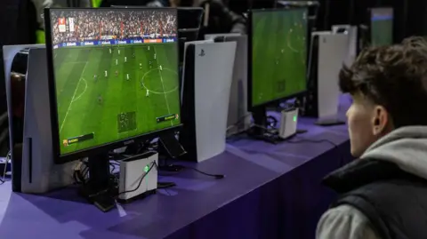 Getty Images A white guy with black hair, wearing a grey hoodie and black gilet sits in front of a small TV playing FIFA next to a Playstation 5.