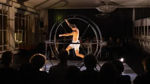 Dorien Hein A topless man, wearing white shorts, stands inside a large ball made of steel hoops. He is striking an acrobatic pose. The ball is on a stage in a darkened room with people watching from all sides.