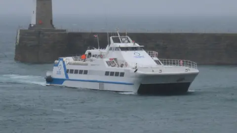 A wide shot of SAN PAWL. It is a large white vessel with a blue stripe running along the side. It is docking in a harbour.