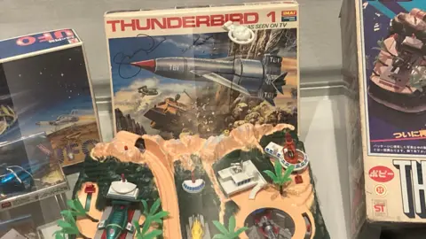 A Vintage Thunderbirds toy playset displayed in a glass case, alongside other boxed models and collectibles.
