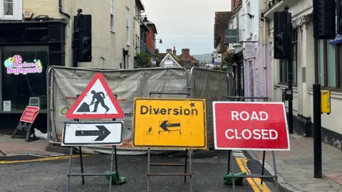 A road closed sign, a roadworks sign and a diversion sign in front of a fence where the sinkhole is