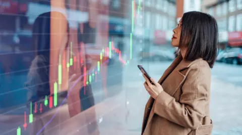 Getty Images A young Asian businesswoman with shoulder length straight black hair and a light brown coat looking at a stock exchange market trading board in the street and checking it against her mobile phone screen