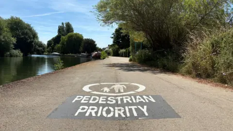 A tow path with a large white 'PEDESTRIAN PRIORITY' sign painted in large white letters on the path. The sign includes a circle with a parent and child painted in the centre. In the background, you can see boats moored.