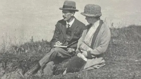 A black and white image of Sydney Bunney and his wife. They are sitting by the coast. He is wearing a hat, dark suit and is sketching in a book. She is also wearing a hat, dress and coat and carrying a handbag. 