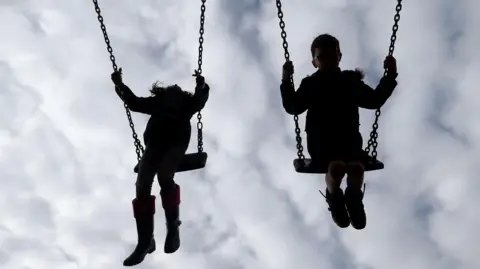 Children on swings. The two children are swinging into the air side by side. Their features are not visible and they appear silhouette-like. It appears to be a young girl on the left and a young boy on the right. The sky behind them is overcast.