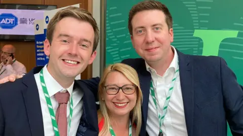 Handout Tom Rutland, Jessica Asato and Liam Conlon smiling as they wear the same green and white striped lanyards
