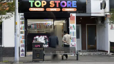 A photo of a mini-mart with a brightly coloured sign which reads Top Store. Outside are ten men with their backs to the camera, leaning against a bench. One is our undercover journalist, which is blurred to protect his identity. The other is the shopkeeper, Surchi.