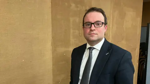 Amy Holmes/BBC Henry Vann in a dark suit and glasses stands indoors against a textured brown wall, with stacked chairs visible in the background.
