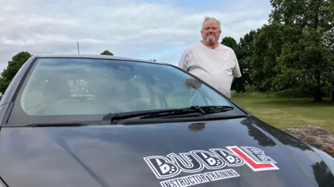 Russell Merchant wearing a white t-shirt and standing beside a black car with his Bubble driving school logo on the hood. In the background there are large trees and a grassy field.
