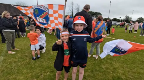 Two boys holding flags standing on a GAA pitch