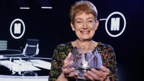Diane Howe with the Mastermind glass bowl trophy. She has short ginger hair and is wearing a black, green and red flowery dress.