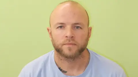 Guernsey Police Kick Bishop - A man with a bald head and a brown beard and moustache wearing a blue t-shirt with a tattoo showing on his neck. he is in front of a green bacground. It is a police mugshot. 
