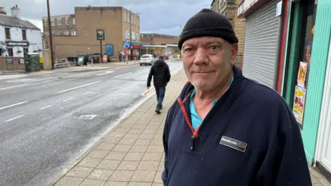 BBC/Elliott Green A man is wearing a black beanie hat and a navy fleece, he is standing in the street