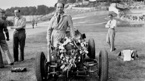National Motor Museum/Heritage Images/Getty Image Uma foto em preto e branco de um homem vestindo um traje de corrida inteiro enquanto se inclina em um carro de corrida ao lado dele com uma guirlanda floral na frente. Alguns homens podem ser vistos ao seu redor em um campo.