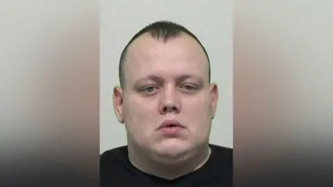 Northumbria Police Mugshot of Beck. He has a large round face and head, is clean shaven and balding with short dark hair which comes to a peak in the centre.