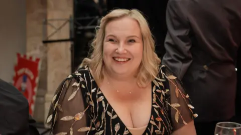 Chloe Docker Chloe Docker sitting at a dinner table with a glass of wine in front of her. She is wearing a short-sleeved black mesh top with gold leaves. She has shoulder-length blonde hair and is smiling.