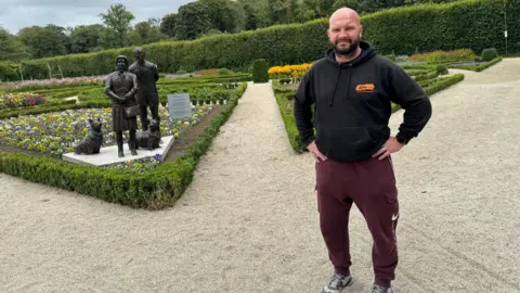 Richard is wearing maroon Nike joggers and a black hoody. He has a black beard and is bald.    He has his hands on his waist and is looking at the camera.