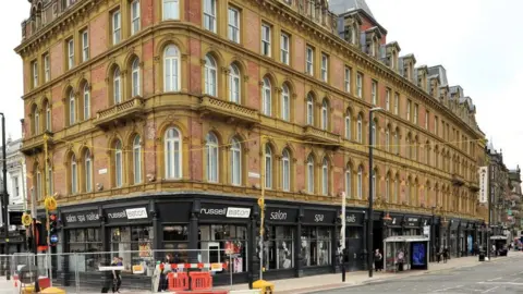 Historic England A 5-storey red brick and sandstone building with large windows. On the bottom floor are shops. There is a bus stop outside and construction work on the corner.