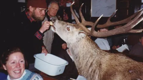 Hostelling Scotland A stag being fed inside Loch Ossian Youth Hostel by a man in a woolly hat, checked shirt and jumper. He is watched by another man, while a woman in the foreground smiles.