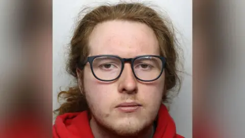 A police mugshot of a sullen man with long, wavy, red hair and black-rimmed glasses. He is wearing a red hoodie.