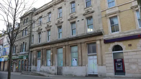 A view of the exterior of the former Lowestoft post office. The yellow brick building's lower windows and doors have been boarded up. The words post office can still be seen above the entrance.