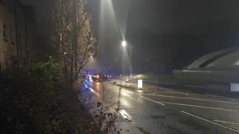 Road lit by streetlights, with a car in the background in the middle of the road with its lights on and a police car in the far distance.