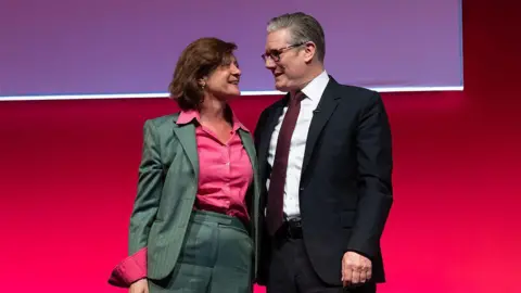 Getty Images Eluned Morgan, in a grey suit and wearing a pink shirt, embraces Sir Keir Starmer, wearing a dark suit, white shirt, glasses and a dark red tie