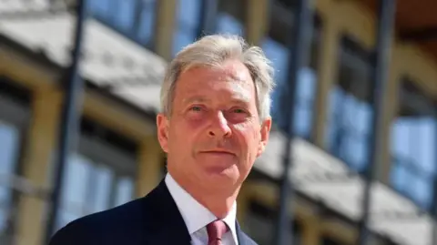 Local Democracy Reporting Service Tim Oliver, wearing a dark suit, white shirt and maroon tie, is pictured standing in front of a modern building on a sunny day.