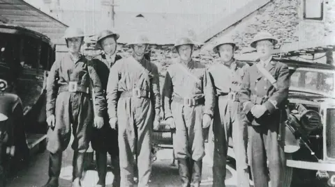 Neil Stanlake Six volunteer firefighters stand together in a black and white photo during World War Two. They are all wearing helmets.