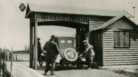 The AA A black-and-white image of the canopied Aldermaston filling station, where a uniformed worker is using a hose to put fuel into a vintage car.