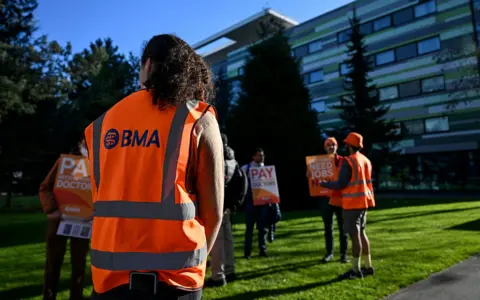 Paul Ellis / AFP via Getty Images A person with their back to the camera wears a hi vis orange vest with the letters 'BMA' on the back, watching as resident doctors stand with placards while picketing outside Manchester university hospital on Tuesday.