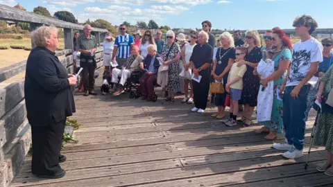 Tim Everest/BBC A group gathering at the Old Shoreham Toll Bridge to mark the 11th anniversary of the Shoreham air crash. A woman in all black is stood at one side, while the congregation is at the other. There are around 25 people in attendance.