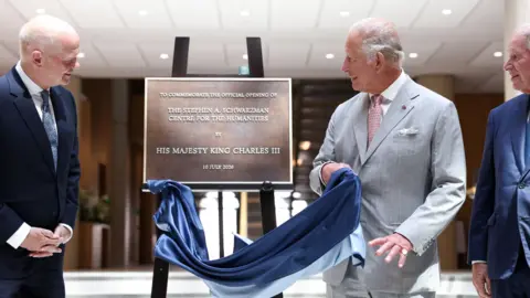King Charles in a grey suit swiping a cloth off a plaque for the Stephen A Schwarzman Centre for the Humanities.