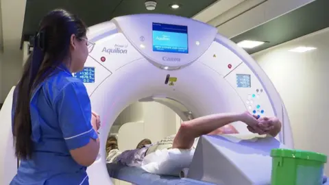 A man lying on his back with his arms outstretched undergoes a CT scan while a female medic with long black hair dressed in a blue uniform in the foreground  watches 