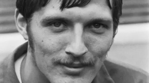 A black and white image of a young man who is a footballer. He has a short fringe, a moustache and is smiling into the camera.