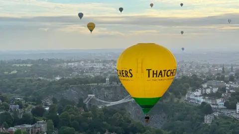 A yellow Thatchers Cider hot air balloon is pictured above the Clifton Suspension Bridge. There are other hot air balloons in the sky in the distance.