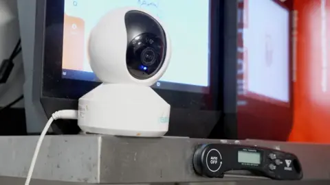 A close up of a white camera on top of a stainless steel shelf. In the background there are screen monitors.
