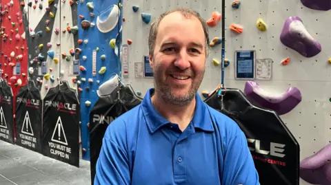 Ollie Conopo/BBC A man with a short beard wearing a blue polo shirt stands in front of climbing equipment. 