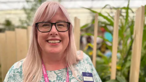 Kathy Grube has blonde and pink hair. She is standing in front of an RHS Chelsea Flower Show exhibition. She is wearing a blue floral outfit, a pair of glasses and is looking at the camera and smiling. 
