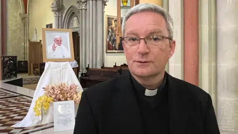 Canon Dominic Golding is sitting in a church in his robes and behind him is a shrine to Pope Francis featuring a photo, flowers and a printed prayer.