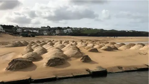 Cormac Mounds of sand piled up on Porth Beach. There are houses on a hill in the background and the sky is cloudy.