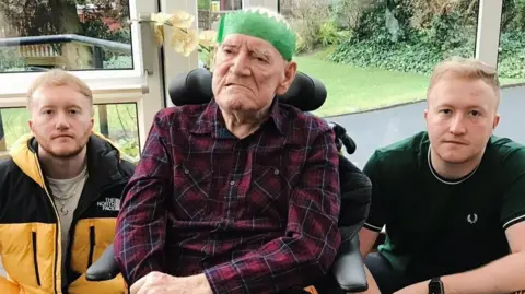 Karen Teasdale-Robson Brian Teasdale sitting in a wheelchair with a headrest, wearing a green paper Christmas party hat and purple and red checked shirt. His twin grandsons - young men - sit on either side of him. Both have sandy coloured hair. One is wearing a green t-shirt and the other is wearing a black and yellow North Face coat.